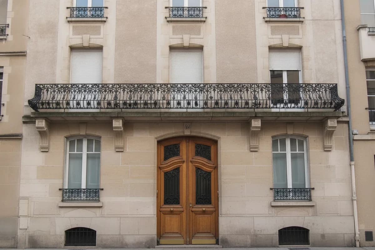 Porte d’entrée en chêne à deux battants vitrés protégés par des grilles, sous un balcon filant en ferronnerie Art nouveau, sur une façade en pierre de taille claire du centre de Nancy